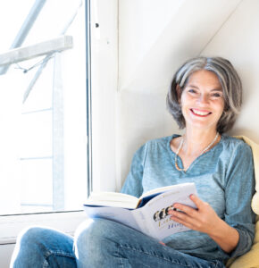 Lerncoach Dagmar Lüdke sitzt lächelnd mit einem Buch in einer gemütlichen Ecke und zeigt Freude am Lernen.